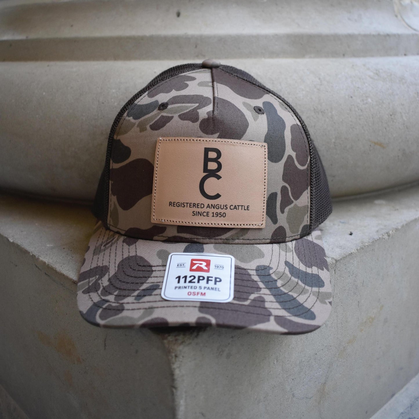 A camouflage trucker cap featuring a tan leather patch on the front with 'B.C REGISTERED ANGUS CATTLE SINCE 1950' and a white 112PFP sticker on the brim.
