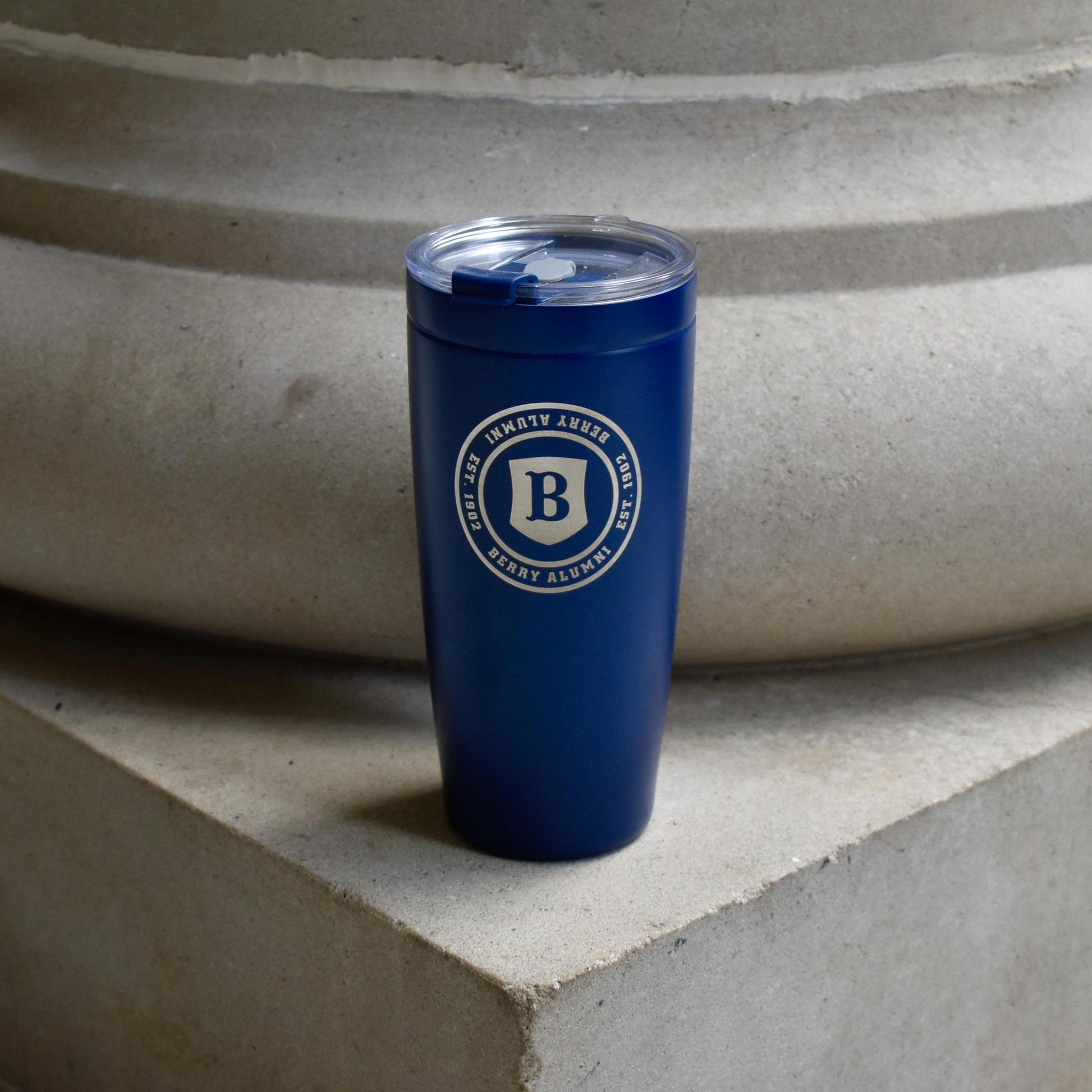 A dark blue insulated tumbler with a clear lid and a circular white logo featuring a large letter B, standing on a concrete ledge in front of a stone structure.